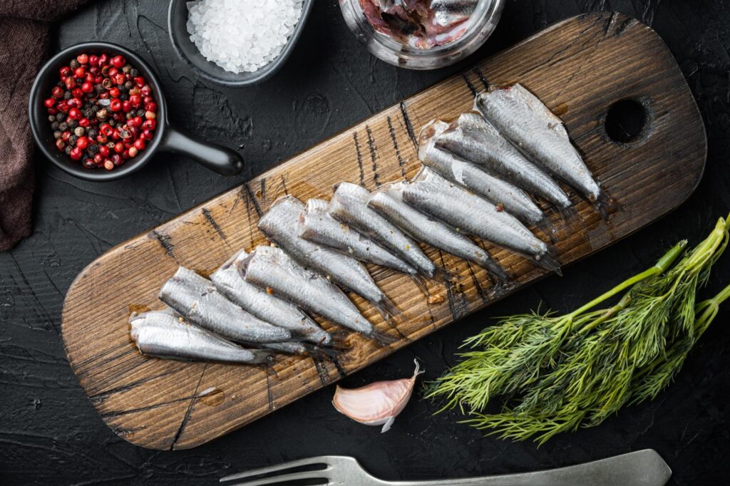 Pickled Anchovy set, on wooden cutting board, on black background with herbs and ingredients, top view flat lay